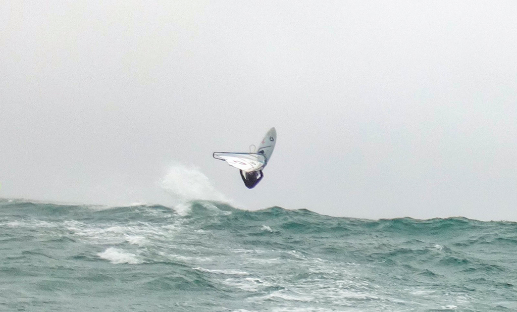 A windsurfer is launched high into the air, performing a jump or trick above a large, churning ocean wave. The windsurfer is captured mid-flight against a hazy, overcast gray sky, with the white sail and board angled upward.